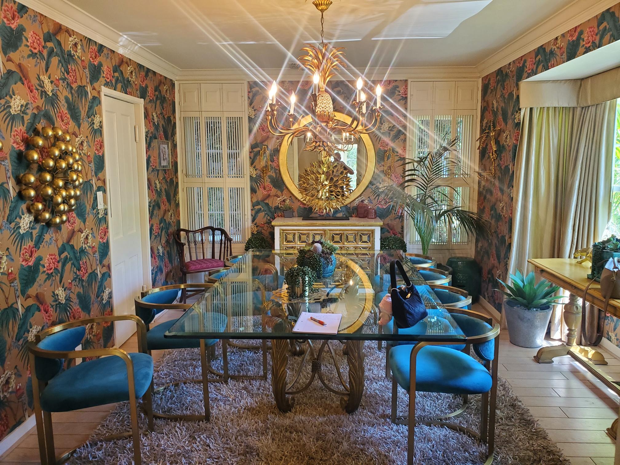 Floral wallpaper dining room with blue velvet chairs, glass table, and gold chandelier